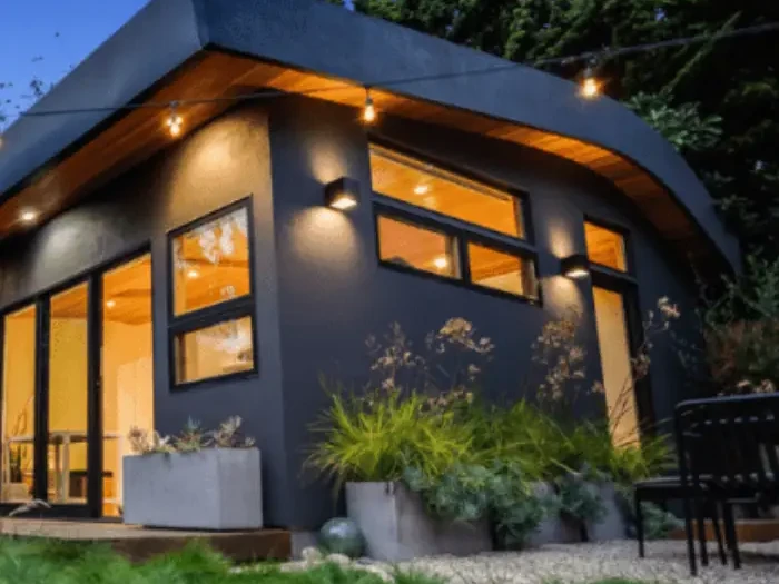 Detached backyard ADU with dark siding, large lit windows and a planted patio at dusk