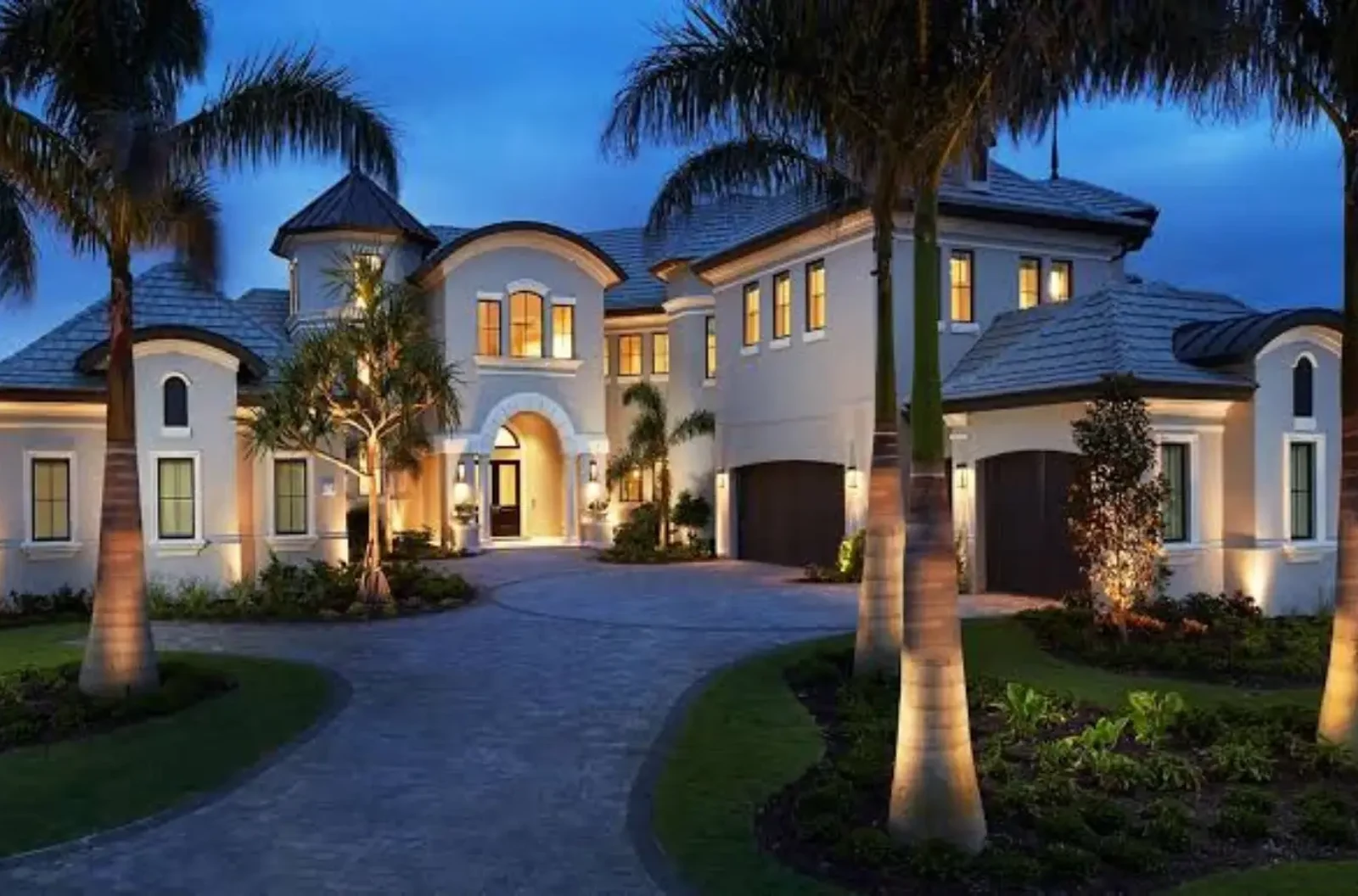 Two-story custom home at dusk with a cream stucco exterior, arched entry, tile roof and a curved paver driveway lined with lit palm trees