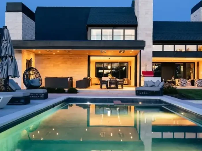 Modern custom home at dusk with a white stone exterior, dark roof, covered patio and a lit swimming pool in the backyard